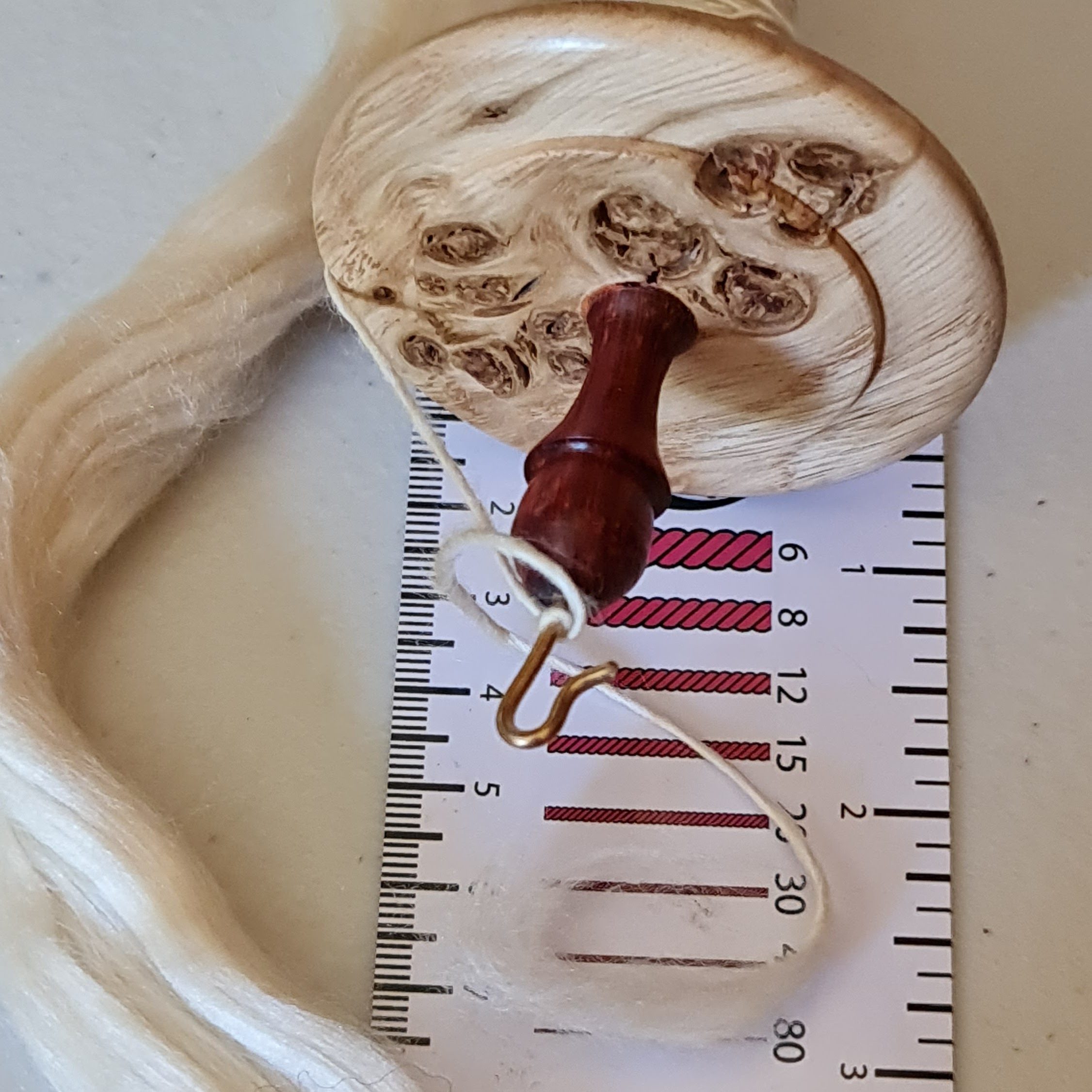 An oak burr spindle with a taupe Seacell single on it, resting on an Electric Eel Yarn Counter card, with some of the unspun fibre in the background. The single is roughly 20 wraps per inch, or about fingering weight.
