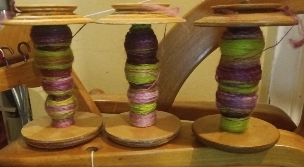 Close-up of 3 bobbins on the integrated Lazy Kate on a Herring Gordon spinning wheel. The singles on the bobbins are bright green and plum.