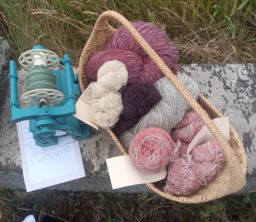 Picture shows a teal Electric Eel Fold 1.0 electric spinning wheel with green yarn on the bobbin, ide-bottomed wicker basket filled with yarn skeins, including a deep pink tweed 2-ply, 2 small skeins in natural white and dark purple, a large natural grey 2-ply skein, and a cake and 2 small skeins of 1-ply in a light coral pink.